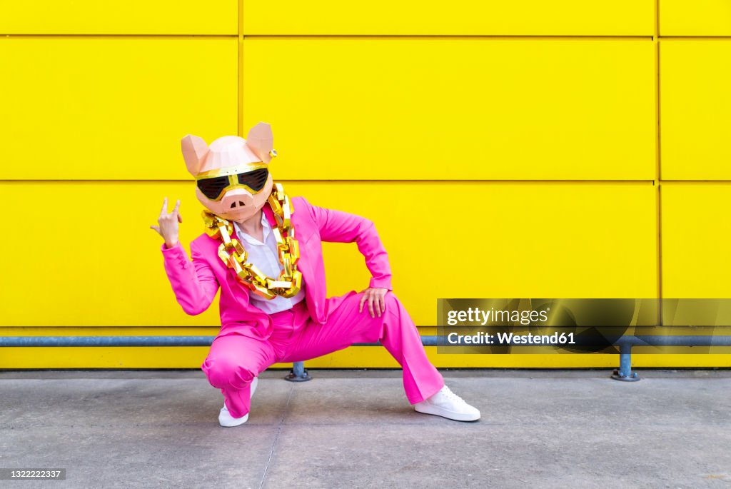 Woman wearing vibrant pink suit, pig mask and large golden chain crouching in front of yellow wall