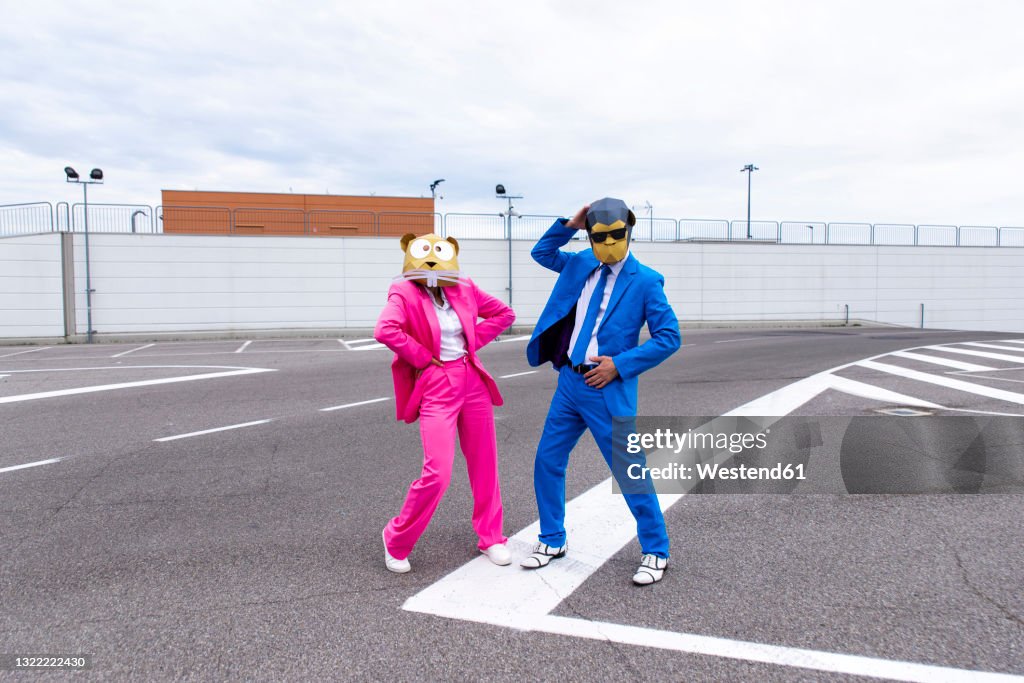 Man and woman wearing vibrant suits and animal masks posing side by side in empty parking lot