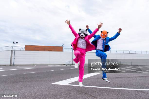 man and woman wearing vibrant suits and animal masks jumping side by side in empty parking lot - animal friends stock-fotos und bilder