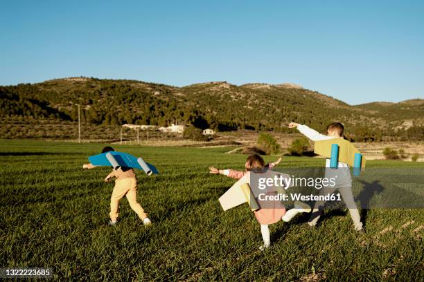 playful brothers and sister playing with rocket wings on agricultural field during sunny day - toy rocket stock pictures, royalty-free photos & images