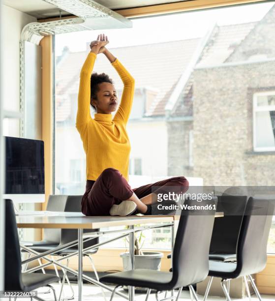 female professional with arms raised meditating on desk at office - office yoga stock pictures, royalty-free photos & images