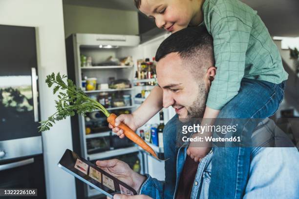 smiling man using tablet while son sitting on shoulder in kitchen at home - smart kitchen fotografías e imágenes de stock