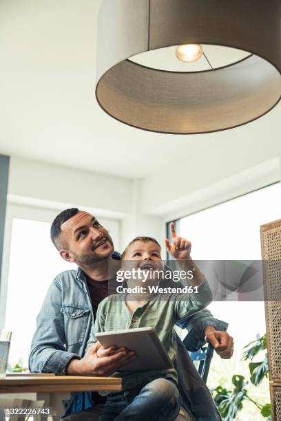 smiling boy pointing at pendant light while sitting with father in smart home - pendant light stockfoto's en -beelden