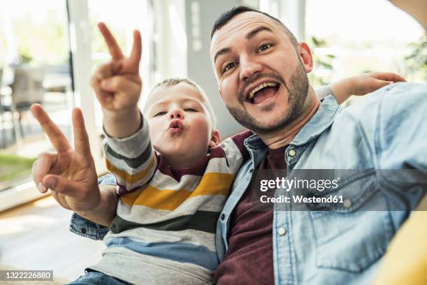 happy father and son gesturing peace sign in living room at home - victory sign stock-fotos und bilder