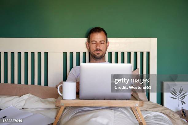 mid adult man napping while lying with laptop on table in bedroom - laziness stock pictures, royalty-free photos & images