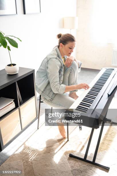 mid adult woman practicing piano while sitting in living room - keyboard white stockfoto's en -beelden