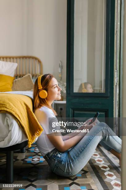 smiling woman looking away while listening music through headphones in bedroom - headphones woman stock pictures, royalty-free photos & images