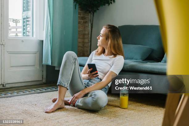 young woman looking away while sitting on carpet at home - barefoot redhead ストックフォトと画像