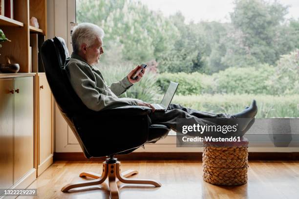 senior man using mobile phone while sitting on chair at home - senior comfortable stock pictures, royalty-free photos & images