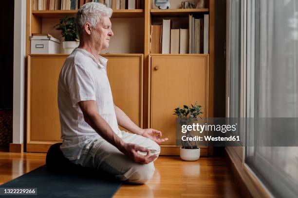 man practicing yoga while sitting in lotus position at home - senior yoga stock-fotos und bilder