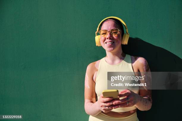 smiling woman with eyeglasses holding smart phone by green wall - woman headphones sport smile iphone stock pictures, royalty-free photos & images