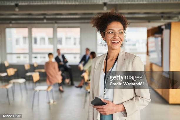 smiling businesswoman with mobile phone looking away at office - black blazer stock pictures, royalty-free photos & images