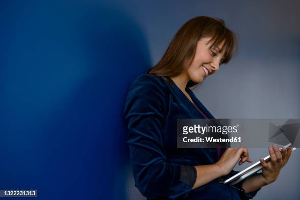 smiling young businesswoman using digital tablet by blue wall in office - blue blazer stock pictures, royalty-free photos & images