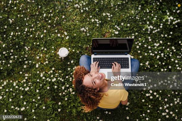 young woman looking up while sitting with laptop on meadow amidst flowers - wiese von oben stock-fotos und bilder