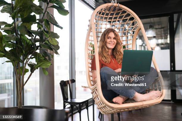 smiling businesswoman with laptop sitting on swing chair at office cafeteria - hanging chair stock pictures, royalty-free photos & images