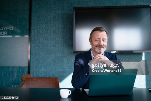 smiling businessman with hands clasped looking at laptop in office - person with in front of screen stockfoto's en -beelden