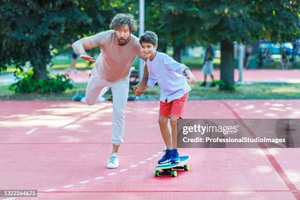 ein junger vater reitet mit seinem sohn in einem öffentlichen park auf einem skateboard. - father longboard stock-fotos und bilder