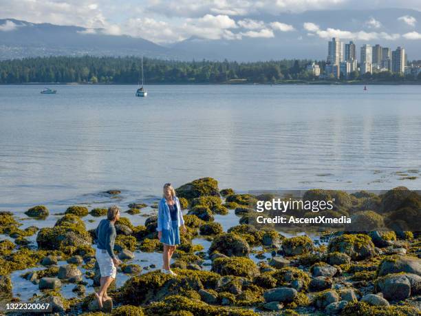 couple cross tidal rocks, city skyline distant - english bay stock pictures, royalty-free photos & images