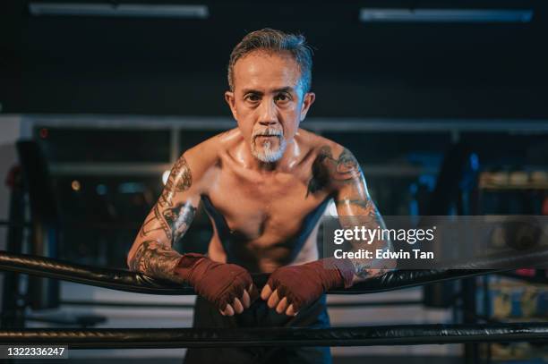asian chinese active senior man boxer with fist looking at camera leaning at side of boxing ring taking a break - muay thai stock pictures, royalty-free photos & images
