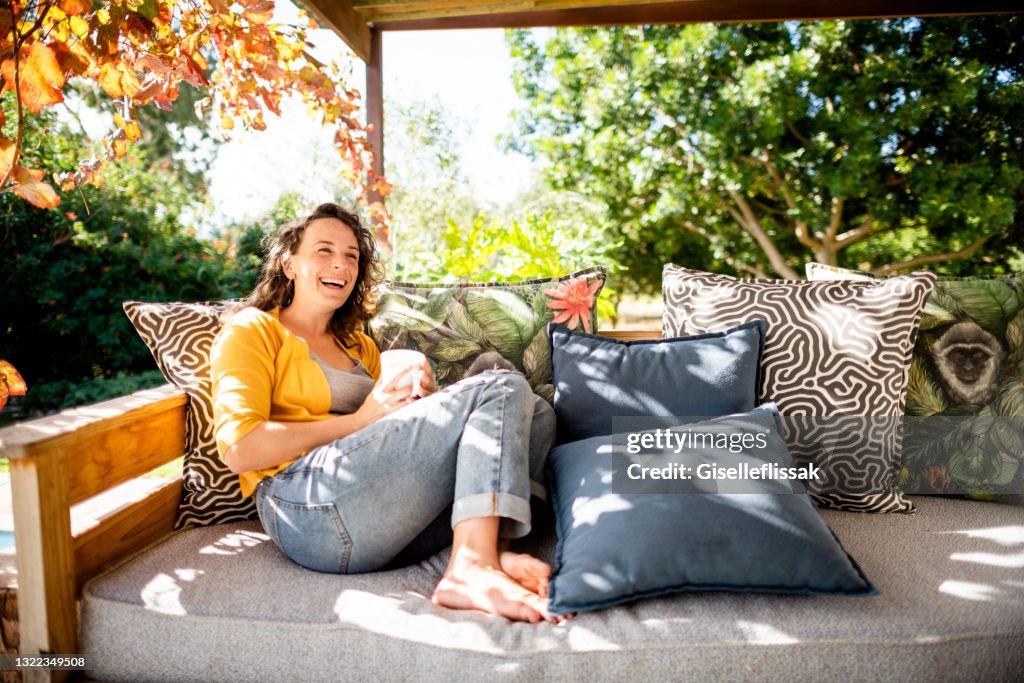 Junge Frau lacht, während sie draußen auf ihrer Terrasse mit einem Kaffee entspannen