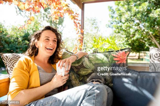 laughing young woman relaxing with coffee outside on her patio sofa - cosy autumn stock pictures, royalty-free photos & images