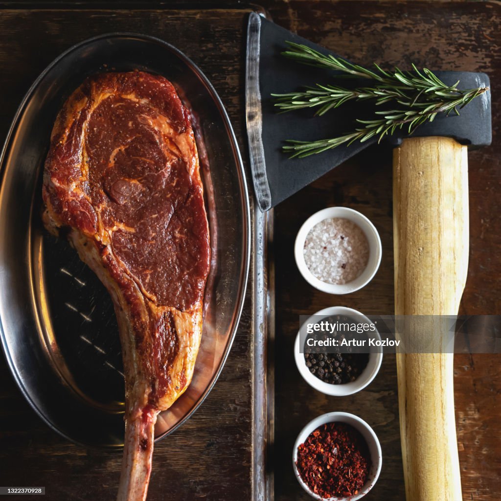 Flat lay, Tomahawk or t-bone steak, cooking process. Butcher ax for cutting meat, large piece of raw meat, and spice set on black background. Top view