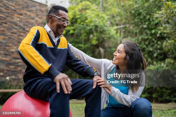 fisioterapista che parla con un uomo anziano seduto su una palla fitness a casa - adults working out foto e immagini stock