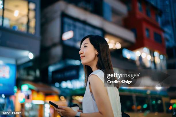 young asian woman walking on the busy downtown city street in the evening, holding a cup of coffee and smartphone on hand, looking away. with illuminated urban city scene and busy traffic in background. lifestyle and technology - hong kong mass transit fotografías e imágenes de stock