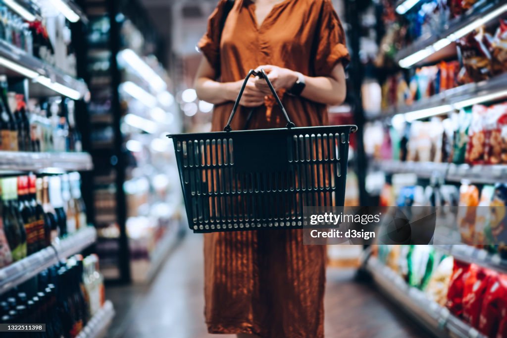 Cropped shot of young woman carrying a shopping basket, standing along the product aisle, grocery shopping for daily necessities in supermarket