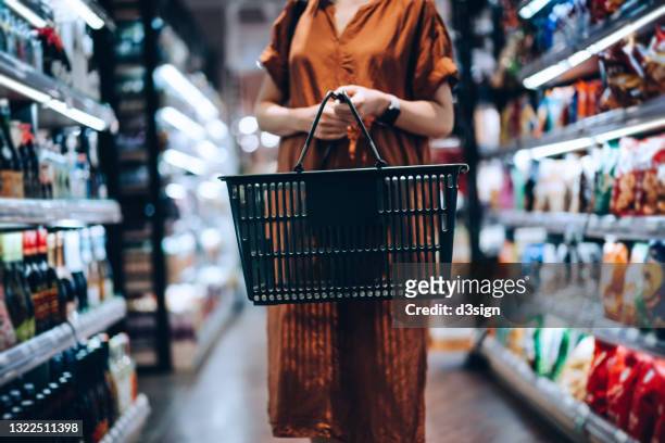 cropped shot of young woman carrying a shopping basket, standing along the product aisle, grocery shopping for daily necessities in supermarket - kaufen stock-fotos und bilder