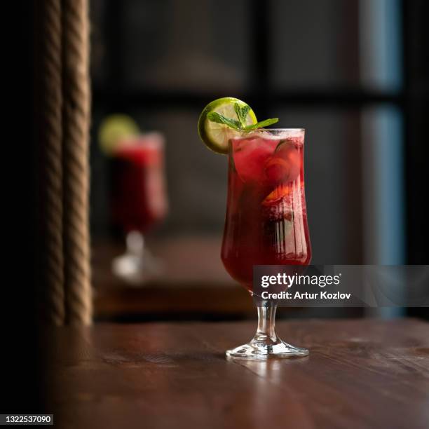 glass of red alcoholic cocktail drink with mint leaf and lime or lemon slice on dark blurred background. side view. copy space - red drink stock pictures, royalty-free photos & images