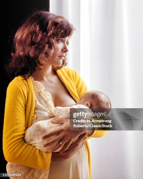 1970s Pensive Brunette Woman Mother Nursing New Born Baby Boy Son Standing Illuminated By Sunlit Window Sheer.