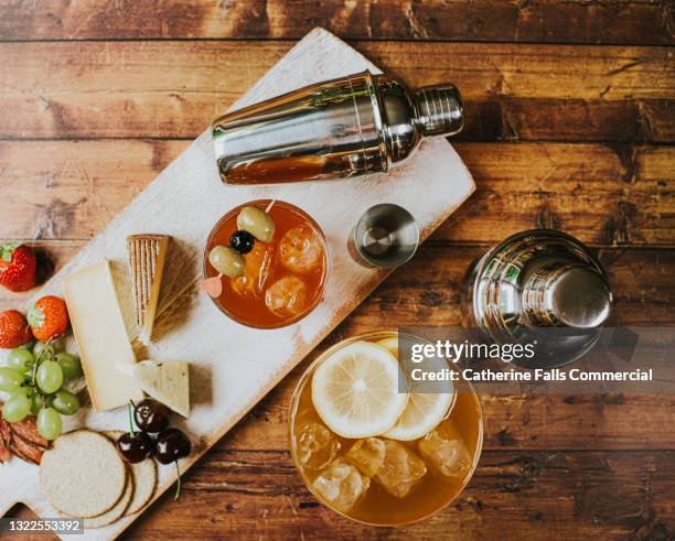 top-down image of a delicious, simple cheese board and a cocktails on a wooden table beside cocktail shakers - spread food fotografías e imágenes de stock