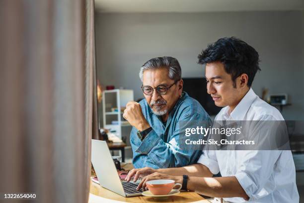 père aîné asiatique et son fils adulte utilisant un ordinateur portable tout en étant assis à la maison - asian discuss photos et images de collection