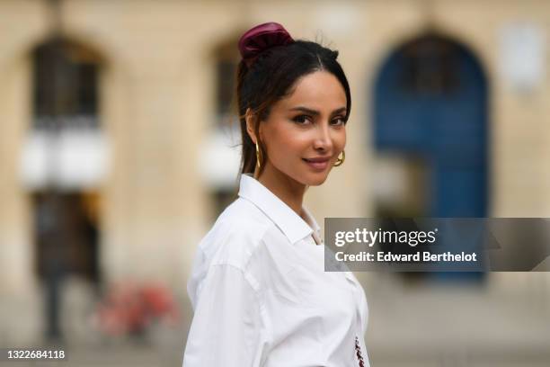 Patricia Contreras wears a burgundy hair elastic, gold earrings / earcuffs, a long white oversized boyfriend shirt, on June 03, 2021 in Paris, France.