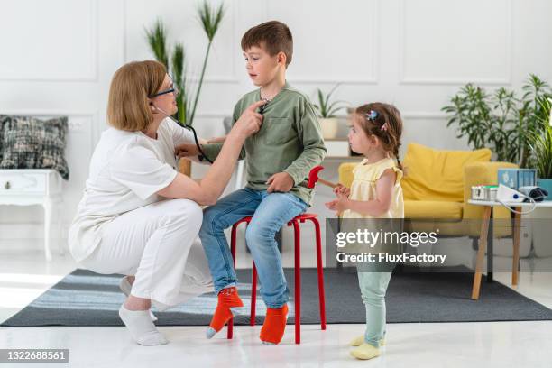 fröhliches kleinkind mädchen, imitieren die kinderärztin, während sie ihren bruder während eines hausbesuchs untersucht - ordination stock-fotos und bilder