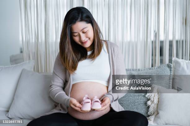 面帶微笑的亞洲年輕孕婦坐在舒適之家的沙發上，肚子上抱著一雙粉紅色的嬰兒鞋。准媽媽期待新的人生觀 - baby booties 個照片及圖片檔