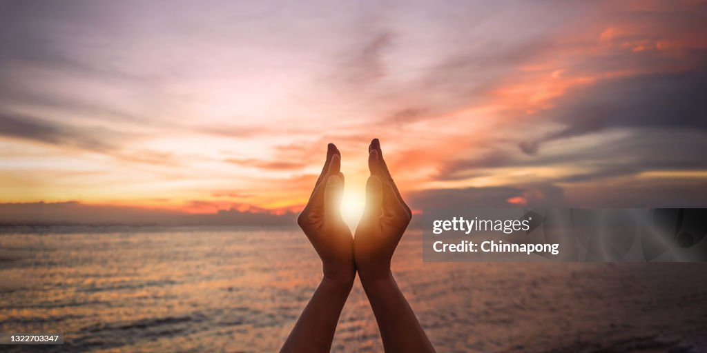 June summer sun solstice concept with silhouette of happy young woman's hands relaxing, meditating and holding sunset against warm golden hour sky on the beach with natural ocean or sea background