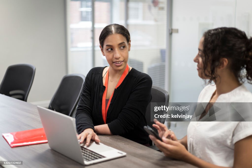 Young woman with a limb difference working on a laptop - looking at and talking to female colleague in office