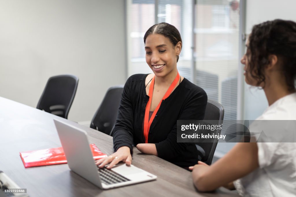 Happy young woman with a limb difference working on a laptop and talking to female colleague in office