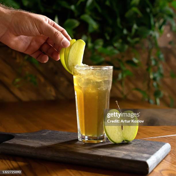 chef decorates apple juice or drink glass with green apple slices. male hand in cooking process of fruit refreshing beverage. vitamins from nature on wooden table, blurred background. soft focus - green apple slices stock pictures, royalty-free photos & images