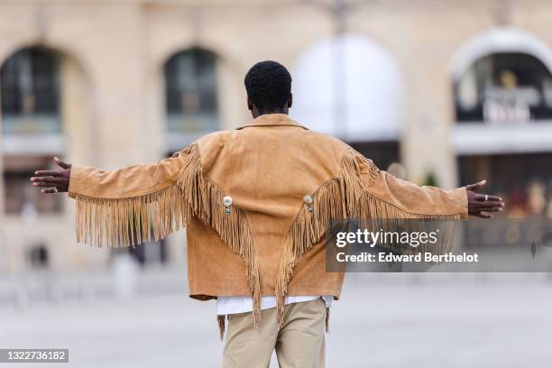 Passerby wears a white t-shirt with black print cross patter, a brown / camel suede fringed oversized jacket with silver an blue pearls buttons,...