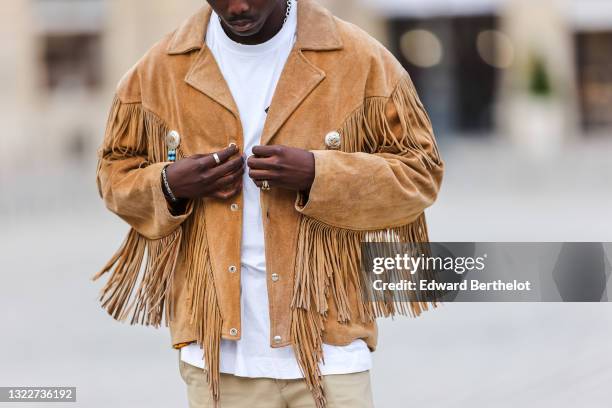 Passerby wears a silver chain necklace, a white t-shirt with black print cross patter, a brown / camel suede fringed oversized jacket with silver an...