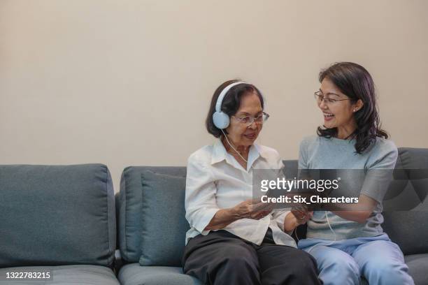 asiatische tochter und ältere mutter, die musik mit kopfhörer und digitalem tablet-computer hört, während sie zu hause auf dem sofa im wohnzimmer sitzt - assisted living stock-fotos und bilder