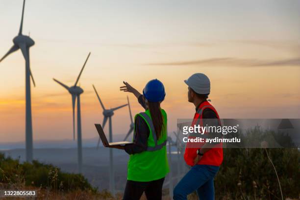 joven equipo de ingenieros de mantenimiento trabajando en un parque de turbinas eólicas al atardecer - windmills fotografías e imágenes de stock