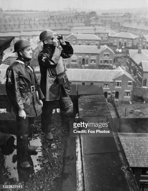Two civil defence firewatchers of the Air Raid Precaution scan the sky with binoculars searching for German Luftwaffe bombers from the rooftop of a...