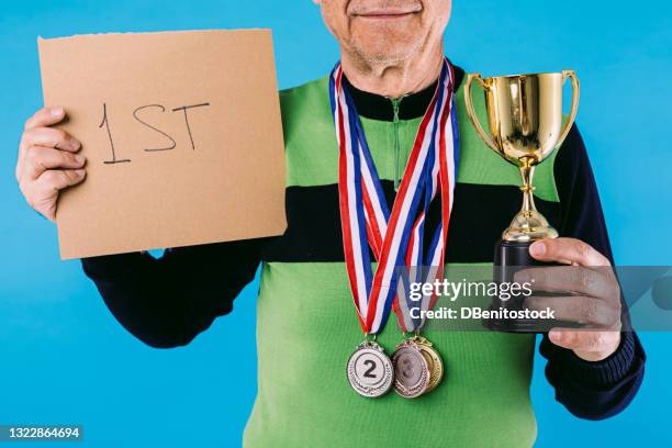 elderly athlete in green and black vintage jersey, with 3 medals hanging, with a sign that reads: "1st" and a trophy cup in the other hand, on a blue background. - first second third place stock pictures, royalty-free photos & images