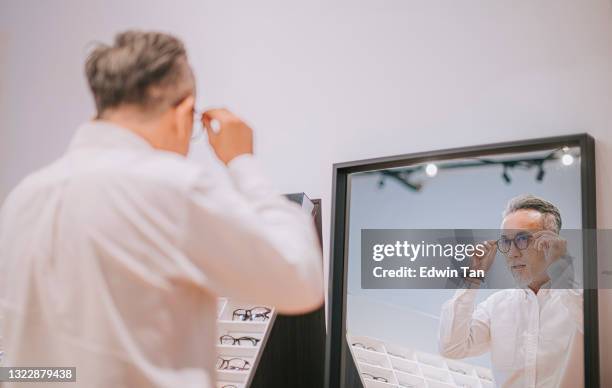 asiatische chinesische senior mann versucht auf brille vor dem spiegel in optiker-shop - optiker stock-fotos und bilder