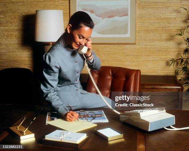 1980s Smiling Business Woman In Office Sitting On Her Desk Talking On Telephone Writing Notes.