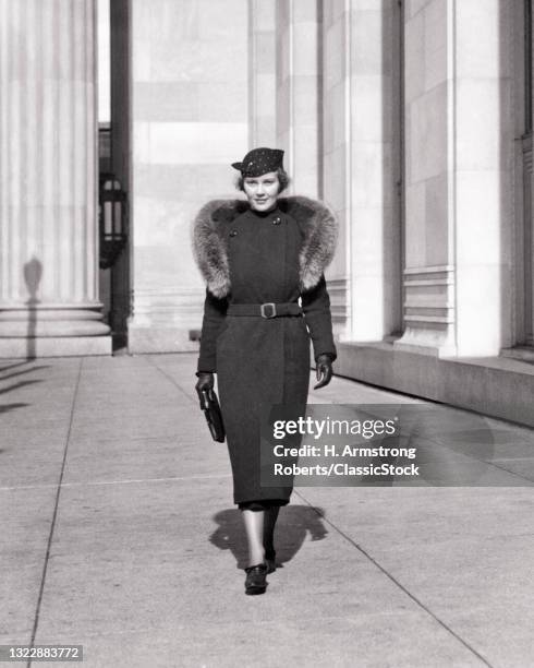 1930s Elegantly Dressed Woman Walking Along Sidewalk Looking At Camera Wearing Belted Fur Trim Coat Hat Gloves Carrying Purse.